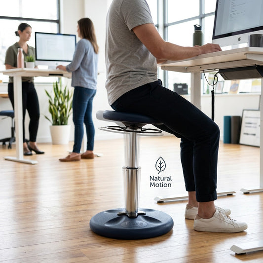 Person using a dark blue fdmt Kore Adjustable Wobble Stool at a desk in a bright office setting.